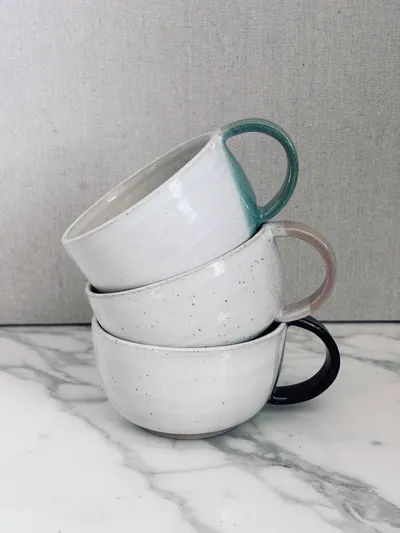 Three stacked mugs with teal, blush, and black handles